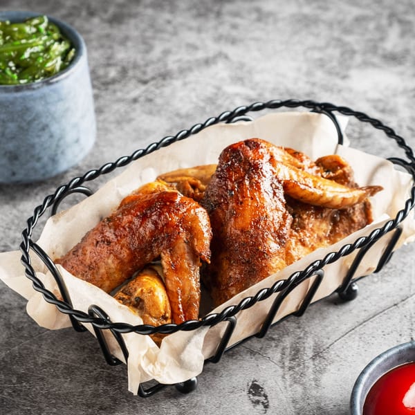 Crispy, glazed chicken wings in a black wire basket with parchment paper, alongside green and red sauces.