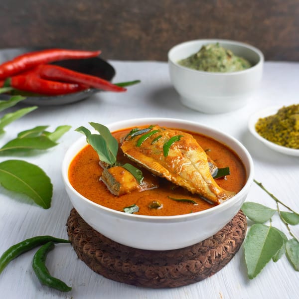 Indian fish curry in a white bowl, garnished with curry leaves, surrounded by spices.